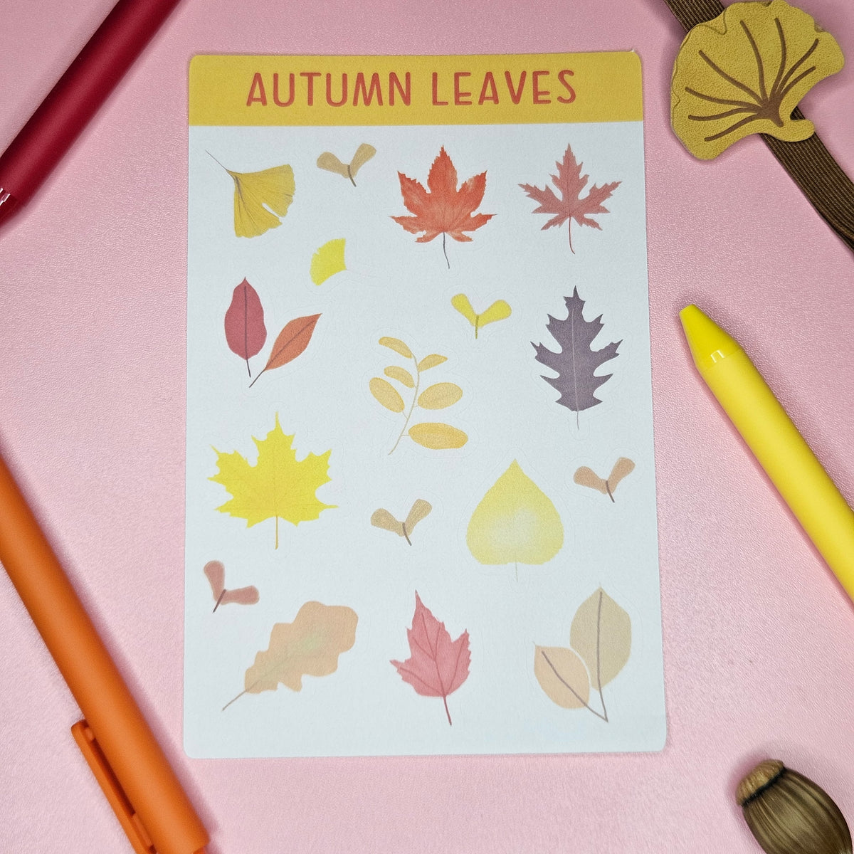 A sticker sheet titled 'AUTUMN LEAVES' featuring various illustrated autumn leaves in yellow, red, orange, and brown. Styled on a pink surface with colorful pens and leaf-shaped accessories.
