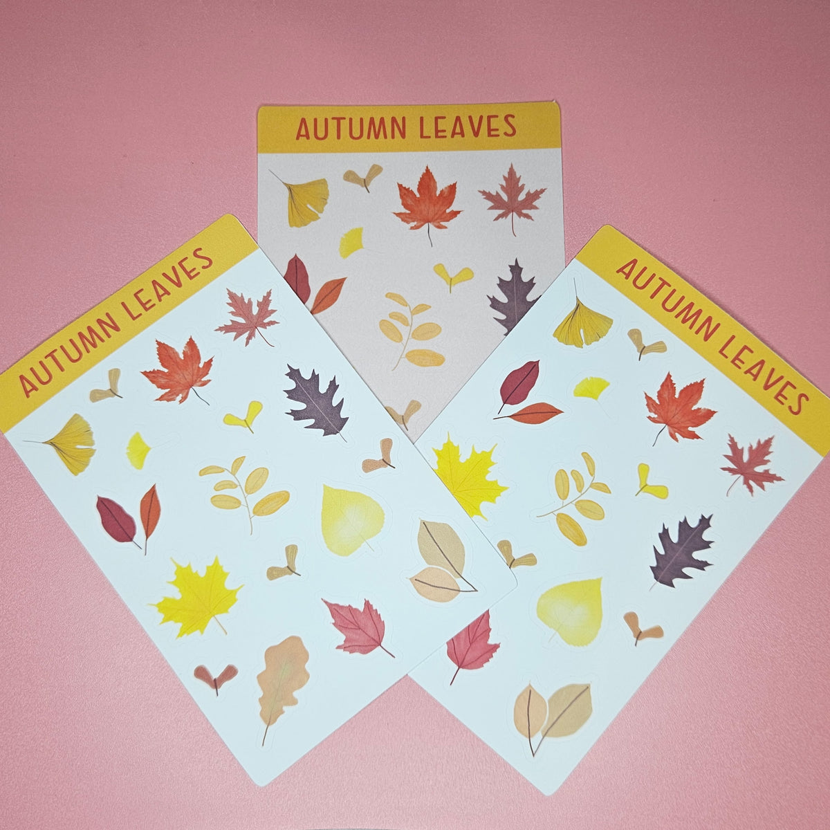 Three sheets of autumn leaf stickers arranged in a fan shape on a solid pink background. The sheets feature various colorful fall leaves in yellow, red, orange, brown, and beige, with a yellow header at the top reading 'AUTUMN LEAVES'.