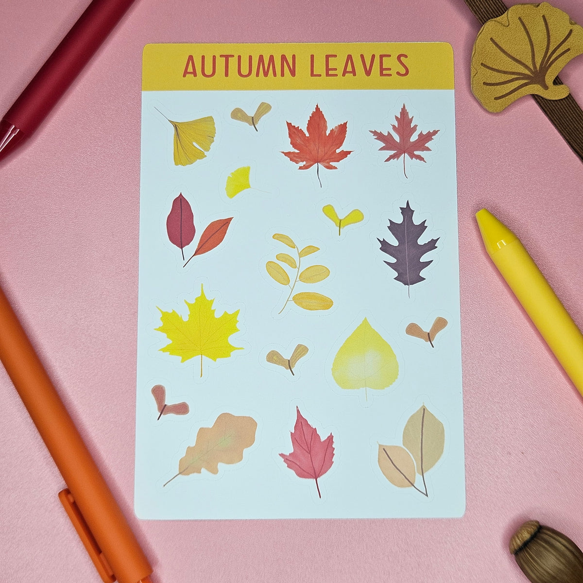 A sticker sheet titled 'AUTUMN LEAVES' featuring various illustrated autumn leaves in yellow, red, orange, and brown. Styled on a pink surface with colorful pens and leaf-shaped accessories.