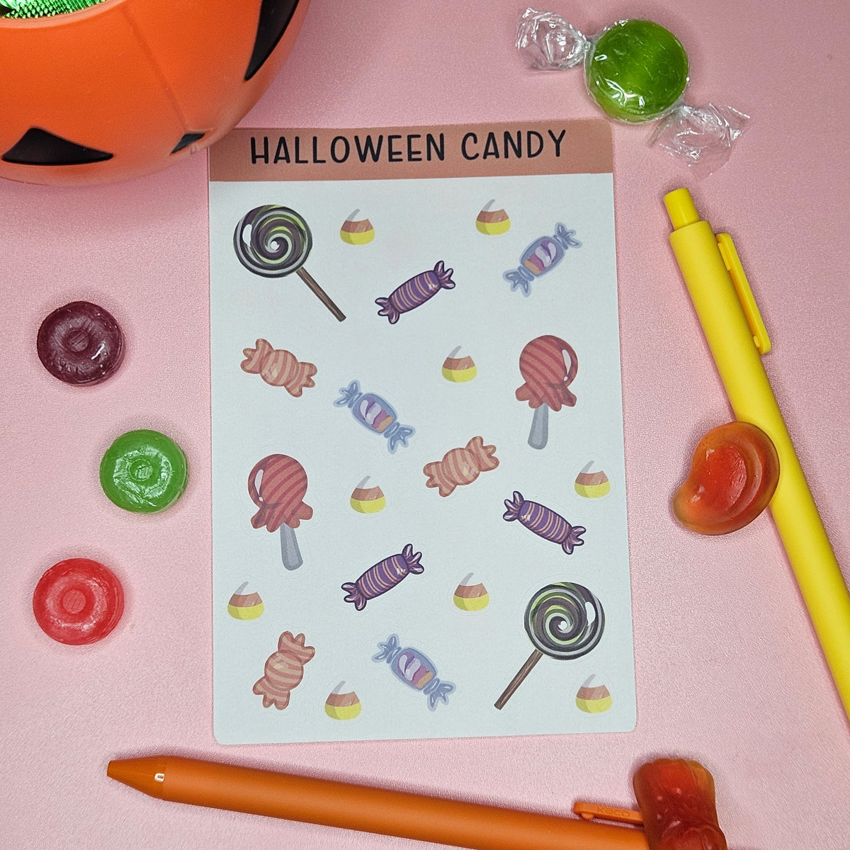 Sticker sheet with a variety of Halloween candy on a pink background with candy and pens around the sticker sheet