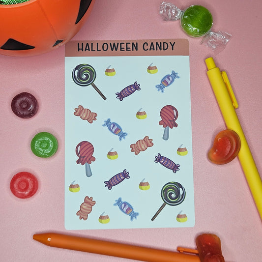 Sticker sheet with a variety of Halloween candy on a pink background with candy and pens around the sticker sheet